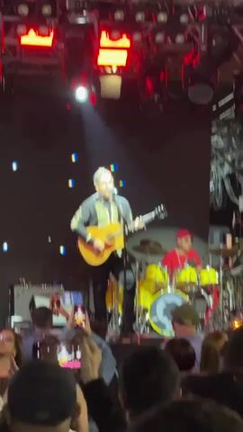This is a video of a live concert performance. In the foreground, the audience is visible, with many people holding up their phones to record the performance. In the background, on stage, a male singer is playing an acoustic guitar and singing into a microphone. He is wearing a dark jacket with gold trim. To his right, a drummer is playing a yellow drum kit. The stage is lit with bright red and white lights, and there are digital displays on the backdrop. The overall atmosphere appears energetic and engaging.
