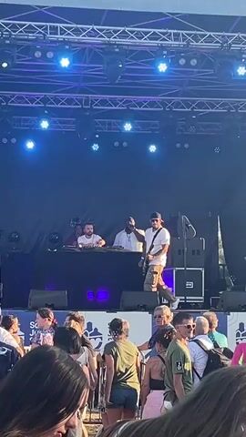 A group of people are on a stage at an outdoor event. In the center of the stage, a person in a white t-shirt, brown shorts, and a hat is playing a guitar. To their left, there are two people behind a DJ booth with turntables and a laptop. To the right of the guitarist, a person in a white t-shirt and dark pants stands. In the background, there are large speakers and stage lighting. In the foreground, a crowd of people are watching the performance. The event appears to be taking place during the day, with a clear sky.