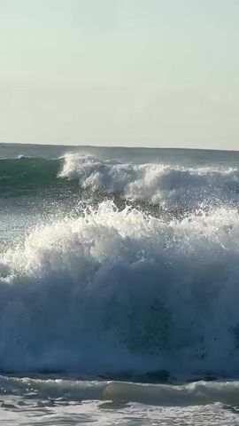 The video shows a dynamic ocean scene. In the foreground, waves are crashing and creating foamy white surf that reaches the viewer's perspective. Further out, larger waves are rolling in, with their crests breaking into white foam. The water is a mixture of blues and greens, reflecting the sky. In the background, the horizon is visible, with a pale, possibly overcast sky above. The overall impression is one of a lively and powerful ocean environment. There are no people or man-made objects visible in the frame.