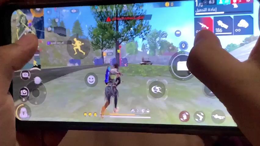 A person is holding a black smartphone horizontally with both hands, actively playing a video game displayed on the screen. The game is a third-person shooter featuring a character in a virtual outdoor environment with trees, structures, and other game elements. The person's thumbs are visible on the lower part of the screen, controlling the game character's movement and actions. The right index finger is briefly visible tapping the top right corner of the screen. The background behind the phone is out of focus, showing indistinct colors.