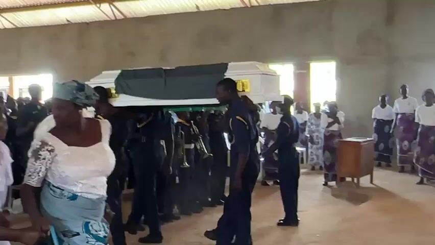 This media file depicts a funeral procession inside a building in Kufana, Nigeria. The central subjects are a group of pallbearers in dark uniforms carrying a white coffin adorned with a green sash. They are walking from left to right across the frame.

Surrounding the pallbearers are mourners. To the left, several women are dressed in brightly colored traditional attire, including headwraps and patterned skirts. Their expressions appear solemn, with some looking towards the coffin and others engaged in conversation. Further back, more individuals are visible, many dressed in white or light-colored clothing. On the right side of the frame, a man in a dark uniform stands facing the procession, possibly ushering or directing.

The setting appears to be a simple, somewhat unfinished building. The walls are bare concrete, and the ceiling is made of corrugated metal. Natural light streams in through several windows, suggesting it is daytime. The floor is covered in dirt or sand. The overall atmosphere is somber and respectful, indicative of a funeral service.

There is no visible text in the media file. The time of day appears to be daytime, and there are no indicators of specific weather conditions. The location context of Kufana, Nigeria, is provided externally. The emotions conveyed are predominantly solemnity and grief.