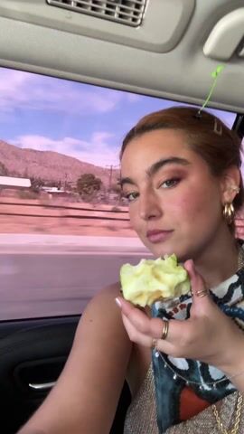 A woman is sitting in the back of a car, holding an apple that has been mostly eaten. She has multiple rings on her fingers and hoop earrings. The background is out of focus, but it appears to be a desert town. The car is moving.  She looks at the apple, and then up and to the side.  She then smiles at the camera. There is music playing.