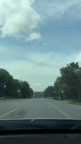 The video shows a view from inside a vehicle driving on a road. The road is paved and has white lines marking the lanes. There are trees and greenery on both sides of the road. The sky is blue with white clouds. There are yellow pedestrian crossing signs visible on both sides of the road. The vehicle passes over a crosswalk with yellow and white stripes. A dark-colored SUV is seen driving ahead of the vehicle. Further down the road, a truck is seen driving towards the camera. A bus stop shelter is visible on the right side of the road.