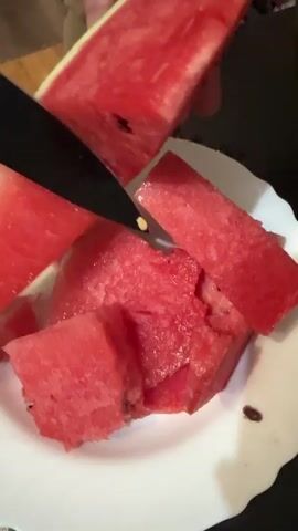 The video shows a close-up view of a watermelon being cut into slices and placed on a white plate. The watermelon is red with black seeds and a green rind. A knife is used to cut the watermelon into small pieces. Some of the watermelon slices are already on the plate, and more are being added. The background is dark and out of focus, but appears to be an indoor setting. There are no people clearly visible in the video, but hands can be seen holding the watermelon and the knife.