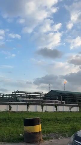 The video shows an industrial complex, possibly an oil refinery or chemical plant, under a partly cloudy sky. A tall chimney emits a constant orange flame.  The plant has a metal framework and a low-rise building with a green roof.  The complex is surrounded by a white wall, with small palm trees lining the perimeter.  A grassy area lies in front of the wall, and a black car is partially visible in the foreground.  Several birds are flying in the sky.