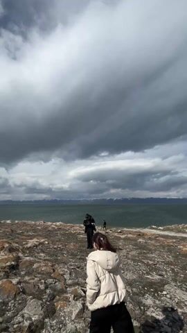 A woman in a white puffer jacket and black pants walks along a rocky shore towards a large body of water. The sky above is dramatic, with heavy, dark grey clouds and some patches of lighter sky. In the distance, snow-capped mountains line the horizon across the water. Another person in a dark jacket stands further down the shore facing the water, and a third figure is visible even further away. The main subject turns to converse with the person in the dark jacket, while the camera captures the expansive, rugged landscape. The scene suggests a cool, cloudy day at a lake in a mountainous region, likely Berdkunk, Armenia.