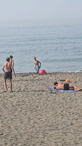 The video shows a sunny beach scene in Málaga, Spain. In the foreground, a person lies on a blue towel on the sand, facing away from the camera, with flip-flops nearby. Further down the beach, three young males are near the water's edge. One stands facing left, while two others are closer to the ocean. One of these two boys is in the process of doing a backflip in the shallow water. A large, bright pink ball is partially submerged in the water, and a smaller orange ball is near the person lying on the towel. The water is calm and reflects the clear sky, suggesting a pleasant, warm day, likely in the afternoon given the sunlight. The overall mood is relaxed and recreational, with people enjoying the beach and the sea. No text is visible in the media.