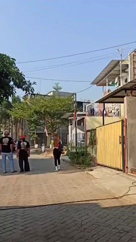 The video captures an outdoor scene with several people present. The environment appears to be a village or residential area, with houses visible in the background and trees providing shade. The ground is paved with brick-like patterns, and a yellow gate is prominent on the right side of the frame. Some of the individuals are walking, while others are standing or sitting on chairs. There are flags and decorations suggesting a festive or community event. A motorcycle is also visible in the distance. The lighting indicates it is daytime, with clear skies.