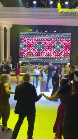 A wedding reception is in full swing with guests dancing in a circle. The room is decorated with elegant columns and the dance floor is lit with purple and pink lights. A large screen behind the dancers displays a vibrant, colorful pattern. In the foreground, several people are holding hands and moving in a circular motion. A woman in a white top with embroidered details and a woman in a white dress are singing. A man in a suit is standing near them. Other guests are observing the dance or mingling in the background. The atmosphere appears festive and celebratory.