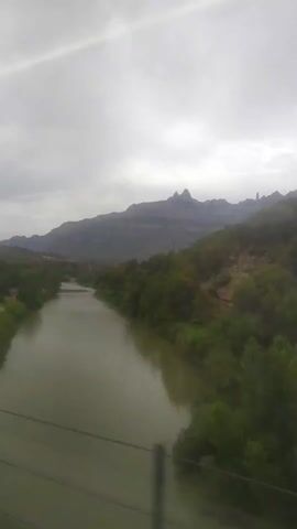 The video captures a wide shot of a river flowing through a valley with mountains in the background. The sky is overcast, with a bright streak of sunlight piercing through the clouds. Lush green trees and vegetation line the banks of the river, creating a vibrant contrast against the muted tones of the mountains and sky. In the distance, the rugged peaks of the mountains are visible, their silhouettes softened by the atmospheric haze. The river itself appears calm and wide, its murky green water reflecting the cloudy sky. The overall impression is one of natural beauty and tranquility.