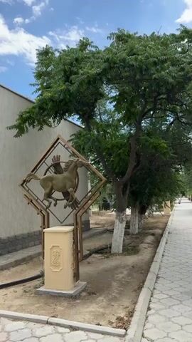 The video shows a paved walkway lined with trees. On the left side of the walkway, there is a decorative metal sculpture featuring a horse with wings and a person. A beige box is positioned below the sculpture. Further down the walkway, a person is seen pushing a stroller. The environment appears to be an outdoor park or garden with a blue sky and scattered clouds.