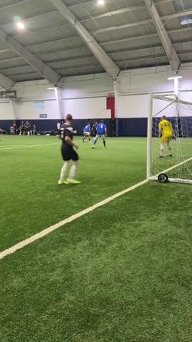 A soccer game is being played indoors on an artificial turf field. A player wearing a black jersey with the number 7 on the back is dribbling the ball. He is facing away from the camera and towards a soccer goal. In the goal, a player wearing a yellow jersey is positioned as the goalkeeper. Several other players in blue jerseys are visible on the field, along with spectators in the background. The environment appears to be a sports complex with a high ceiling and overhead lighting.