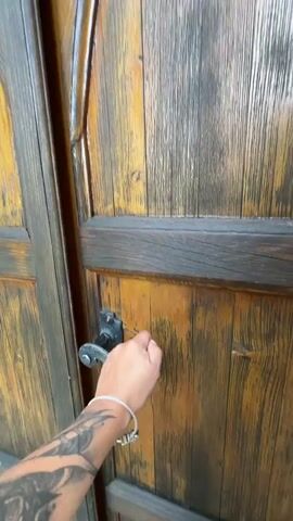 The video begins with a close-up of a person's hand, which has tattoos on the forearm and a bracelet on the wrist, reaching for a dark metal door handle on a large, weathered wooden door. The door then opens, revealing the interior of a small chapel. The camera moves forward into the chapel, showing a simple wooden bench on the left side and another on the right. In the center, there is a small altar area enclosed by a decorative metal gate. Behind the gate, on the blue-painted wall, is a colorful painting of several women with diverse skin tones. The altar itself is adorned with various small religious statues, framed pictures, and potted plants. A stained-glass window is visible on the left wall. The floor is tiled. The camera continues to move slowly towards the altar area, focusing on the details behind the gate.