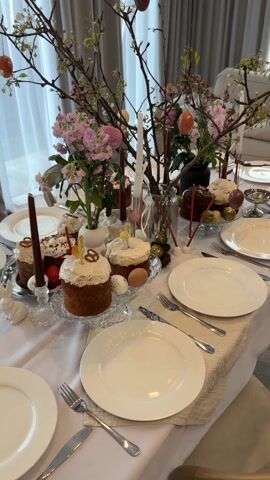 The video showcases a beautifully decorated table set for an Easter celebration. The table is covered with a white tablecloth featuring a runner with printed designs. Place settings are arranged for multiple people with white plates, silverware, and napkins. Several Easter cakes are displayed, some with white icing and decorations, while others are chocolate. There are also colored eggs, some plain and some painted, placed around the cakes. Vases of fresh flowers, including pink stock, and decorative branches with hanging Easter eggs are interspersed among the food. Candles in candlesticks and small decorative nests with golden eggs add to the festive atmosphere. The background features sheer curtains, suggesting the setting is indoors, likely a dining room or living room. The overall impression is of a meticulously arranged and elegant Easter feast.