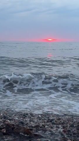 The video shows a beach with a calm sea and a sunset in the distance. The waves are gently crashing on the shore, which is covered in pebbles. The sky is a soft gradient of pink and grey, and the sun appears as a bright red orb just above the horizon, casting a shimmering reflection on the water. The scene is peaceful and serene.