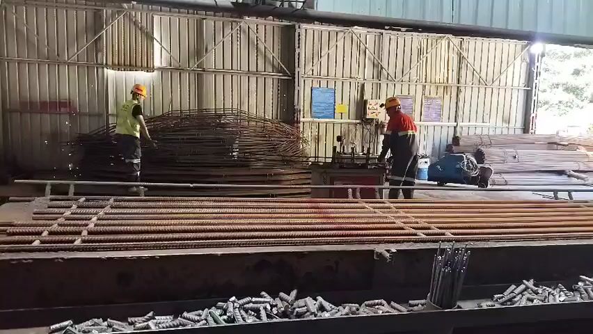 This video shows a construction or manufacturing setting, likely a steel fabrication workshop. In the foreground, rows of rebar (reinforcing bar) are laid out on a metal surface. Further back, several individuals are working. On the left, one person in a yellow hard hat and a high-visibility vest is near a large pile of bundled rebar. In the center, two individuals, both wearing hard hats and high-visibility vests with red and orange accents, are operating machinery. One person is positioned at a machine that appears to be bending or cutting rebar, with sparks visible, suggesting cutting activity. The other person is standing near a pile of wire mesh or cages. To the right, a blue industrial machine is visible, and beyond that, stacks of long metal rods. The environment is a large, open-air structure with corrugated metal walls and roof. The lighting suggests it is daytime, with natural light entering from the open sides. The overall impression is one of industrial activity involving metal reinforcement materials.