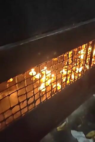 The video shows a close-up view of a grid structure, possibly a fence or grate, behind which numerous small oil lamps are lit. The flames of the lamps are visible through the openings of the grid, casting a warm, flickering glow. The lamps appear to be made of metal and are arranged in rows, suggesting they are part of a display or ritual. The background is dark, emphasizing the illumination provided by the lamps. The camera slowly pans across the scene, revealing the density and arrangement of the lit lamps. There are no people visible in the video, and the focus is solely on the lamps and the grid.