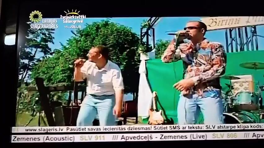 The video shows two men singing on an outdoor stage. The man on the left wears a white short-sleeved shirt and blue jeans, holding a microphone and singing. The man on the right wears a colorful patterned shirt and blue jeans, also holding a microphone and singing. There is a green backdrop behind them, along with some trees and foliage. In the lower part of the screen, there is text overlay with a website address and some song titles. The overall scene appears to be a live music performance.