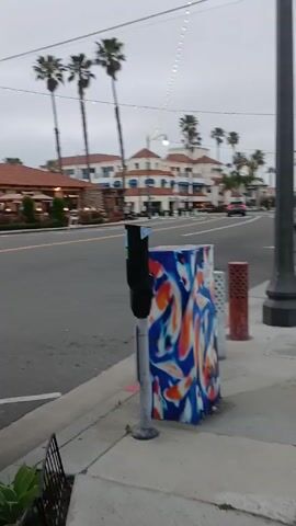 The video shows a street scene in a coastal town. The sky is overcast. There are palm trees lining the street. On the left is a colorful utility box with a koi fish design. There is a parking meter next to it. In the distance are buildings with Spanish-style architecture. A "Road Work Ahead" sign is visible on the sidewalk. A few people are walking in the distance. A car drives down the street. The camera pans slowly to the right.