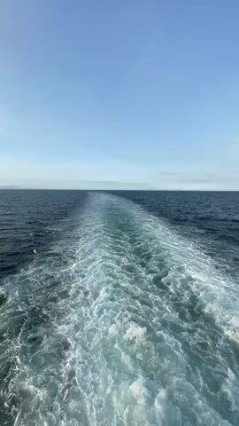 The video shows the wake of a boat or ship traveling on a body of water, presumably the ocean, under a bright blue sky with scattered clouds near the horizon. The wake is prominent, characterized by frothy white water contrasting with the darker blue of the surrounding sea. The camera angle is from the rear of the vessel, looking out towards the horizon. There is a white railing visible on the side of the vessel. In the distance, a faint landmass can be seen.