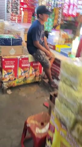 A blurry, handheld video captures a young person wearing a dark t-shirt, camouflage shorts, a dark baseball cap, and sandals, sitting on a counter in what appears to be a store or market. They are looking to their right. The environment is filled with stacked merchandise, including boxes and large packages of what looks like instant noodles or snacks. Behind the person, shelves are stocked with various brightly colored items. In the foreground, a red stool is visible, and more product packaging is stacked on the right side of the frame. The overall scene suggests a busy retail setting, though the focus is on the seated individual.