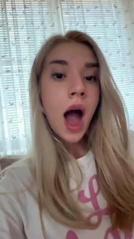 A blonde woman with her hair down speaks directly to the camera. She is wearing a white t-shirt with pink lettering. Her facial expression changes from concerned to surprised to annoyed. The background is a window with blinds.