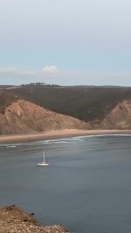 A sailboat is visible on the water near a beach. The beach is bordered by steep cliffs. The sky is overcast.