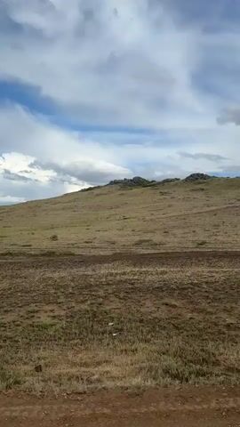 The video captures a scenic view of a vast, rolling landscape in Bakty, Kazakhstan. The terrain is characterized by dry, grassy hills with patches of green vegetation, suggesting a semi-arid environment. A dirt road runs horizontally across the lower portion of the frame, indicating it's a rural or undeveloped area.

The sky is dramatic, filled with a mix of white and gray clouds against a backdrop of blue sky, hinting at dynamic weather conditions, possibly with the potential for rain or a recent shower. The time of day appears to be overcast daylight, with diffused light across the landscape. There are no people or discernible objects other than the natural elements and the road.

The overall impression is one of open space and natural beauty, with a slightly rugged and untamed feel. The location cues, such as the dry grass and the type of terrain, are consistent with a steppe or grassland environment. There is no visible text in the video. The lack of human presence emphasizes the scale and solitude of the natural setting.