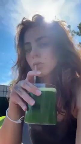 The video shows a woman with brown hair drinking a green beverage with a straw from a glass. She is outside, possibly at a cafe or restaurant with a view of a lake or river. The sun is shining brightly. She looks directly at the camera while drinking. The camera pans slightly to show the view behind her.