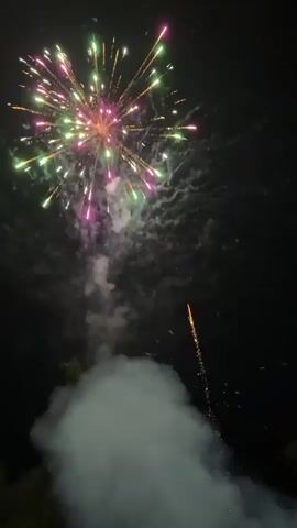 The video captures a nighttime display of fireworks. The first few seconds show a single burst of white sparks against a black sky, accompanied by a plume of white smoke rising from below. Then, a more elaborate burst of colorful fireworks explodes, featuring red, green, and pink sparks radiating outwards, all while the smoke continues to rise. The sequence repeats with a slightly different pattern of colorful bursts and smoke. The scene appears to be outdoors, with dark silhouettes of trees visible along the bottom edge of the frame in some shots, suggesting the fireworks are being launched from ground level.