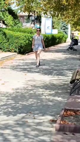 A woman wearing sunglasses, a white t-shirt, and denim shorts walks down a paved path lined with trees and bushes. She is holding sunglasses in her left hand. A stroller is visible in the background, and a bench is on the right side of the path. The sun is shining, casting shadows on the ground.