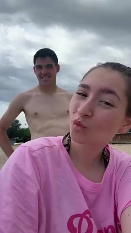A woman and a man are on a beach in cloudy weather. The woman is in the foreground, wearing a pink shirt with a graphic and posing with a pouting face or closed eyes. The man is in the background, shirtless, flexing his arms, and smiling. The beach is sandy, and there are trees and a small building visible in the distance. The sky is overcast.