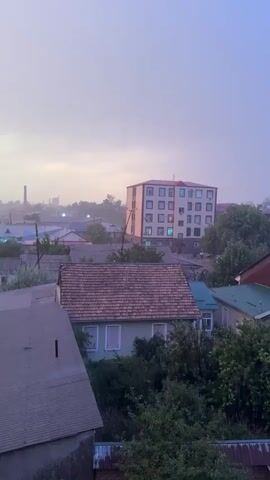 This is a vertical, shaky video of a suburban landscape during what appears to be a rain shower. The sky is overcast with a purple-gray hue. In the foreground, a grey roof of a building is visible. Beyond that, a light green house with a brown tiled roof is situated amongst dense green foliage. In the background, a multi-story building with many windows is illuminated from within by several glowing lights. The overall atmosphere is muted and damp. There are no people or discernible actions occurring. The video is very short, lasting only a few seconds.