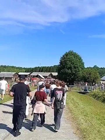 The video shows a group of people walking down a dirt path towards a festival or event. There are wooden buildings and trees in the background, and a stage with lights and speakers is visible in the distance. The sky is blue and clear. Some of the people in the foreground are wearing casual clothing, while others appear to be in traditional attire. The overall atmosphere seems to be festive and lively.