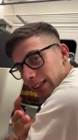 A close-up video shows a man with short brown hair, wearing black-rimmed glasses, and a white t-shirt. He has a small blue stud in his nostril and a piercing in his ear. He is looking at the camera while making a gesture with his hand, extending his index finger and pinky. The background is slightly blurred, but it appears to be the interior of a vehicle, possibly a train. There are other seats visible and some overhead compartments. There is also background noise.