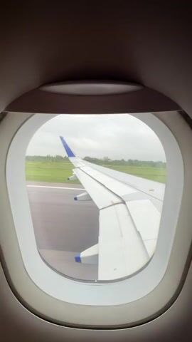 The video shows a view from an airplane window as the aircraft taxis on a runway. The right wing of the plane is visible, extending across the frame. The runway is dark asphalt, with white lines marking its edges. Beyond the runway, there are green fields and a cloudy sky. The plane appears to be moving slowly.
