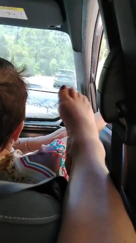 The video shows a person's foot resting on a car seat, with a child's head visible in the foreground. The child appears to be looking out the window at other cars on a road. The interior of the car is dimly lit, with sunlight filtering in through the windows. The child is wearing a colorful outfit. The overall impression is one of a family traveling in a car.