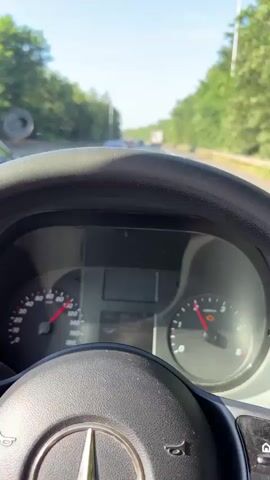 The video shows a first-person perspective from inside a vehicle, driving on a highway. The dashboard displays the speedometer and tachometer, with the needle on the speedometer indicating a speed of approximately 120. The tachometer shows the engine RPM around 2. The view outside the windshield reveals a multi-lane highway with other vehicles. In the foreground, the steering wheel is visible. Trees and vegetation line both sides of the highway, creating a green corridor. The sky is clear and blue, suggesting a sunny day. Several cars are driving in the same direction, and some are in adjacent lanes. A truck is visible in the distance. The road appears to be well-maintained, with clear lane markings. The overall scene depicts a typical highway driving experience on a clear day.