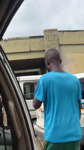 The video captures a street scene from inside a vehicle, looking outwards. The focus is on a man in a turquoise t-shirt and green shorts standing outside a white minivan. He appears to be interacting with the driver or a passenger of the minivan, as his body is angled towards the open rear door of the vehicle. Another person, wearing a red cap, is partially visible inside the minivan. The minivan itself is positioned behind another white vehicle, suggesting a traffic jam or parked vehicles. In the background, there is an overhead structure, possibly a bridge or overpass, with a distinct textured pattern on its pillars and underside. The overall environment appears to be an urban or semi-urban setting with visible debris or uneven ground. The camera perspective from within the car, with a car door and interior components visible in the foreground, suggests the filming person is a passenger or the driver. The lighting indicates an overcast or cloudy day.