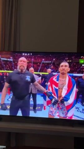A screen displays a live broadcast of a UFC event. On the left, a muscular referee with a beard and tattoos stands facing the camera. He wears a black shirt with a UFC logo on the left breast and dark pants. To his right, a fighter stands wearing a British flag draped over his shoulders and patterned shorts. He has numerous tattoos. A man in a sparkly tuxedo stands between the referee and the fighter, holding a piece of paper and speaking into a microphone. The fighter raises his arms, holding the flag, and appears to be celebrating. The background shows a crowd of spectators and the arena environment.