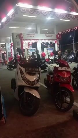 The video shows a gas station at night, decorated with red and white streamers. Two scooters are parked in the foreground, a white one on the left and a red one on the right. In the background, several people are visible, some appearing to be refueling their motorcycles. There are also several other motorcycles parked in the background. The gas pumps are lit and appear to be in operation. The overall impression is of a busy gas station during a night-time event or promotion.
