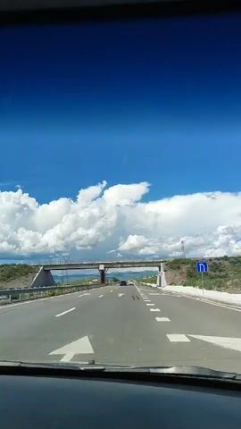 This is a first-person perspective video of someone driving on a highway. The driver is traveling on a multi-lane highway under a bright blue sky with large white clouds. An overpass is visible in the distance, and the car passes under it. Other vehicles can be seen on the highway, including a white SUV passing on the left. The landscape on the right side of the highway is hilly and covered with vegetation. The video captures the experience of driving on a sunny day with dynamic cloud formations.