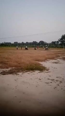 The video shows an outdoor scene, possibly a sports field, with several individuals and motorcycles present. The ground appears to be a mix of dry grass and sandy soil. In the background, there are trees and utility poles with wires. The sky is overcast. Several people are scattered across the field. Two people are on motorcycles, one appears to be stationary, and the other is moving. Other individuals are standing or walking. The overall impression is of an informal gathering or activity taking place in an open area.