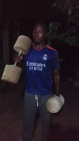 A young Black man wearing a blue and white t-shirt and gray sweatpants is standing outdoors in the dark, holding a dumbbell in each hand. The dumbbells appear to be homemade, with wooden weights attached to metal handles. The man is looking directly at the camera and appears to be sweating. The background is dark and out of focus, with some foliage visible. The man is performing a bicep curl exercise, raising one dumbbell at a time towards his shoulder.