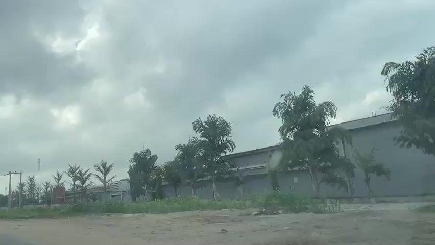 A cloudy, overcast day. The video shows a road with some buildings and trees alongside it. A few people on motorcycles can be seen, as well as some parked vehicles. The road appears to be in a somewhat rural or industrial area. The sky is heavily clouded, with dark clouds suggesting the possibility of rain. The greenery alongside the road is somewhat sparse and dry in some areas, while other areas are more lush. The buildings appear to be industrial or commercial in nature, with large, grey structures. There are also power lines visible, indicating that this is a populated area.