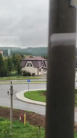 The video captures a view of a residential area on an overcast day. In the foreground, there is a grassy embankment with some brown soil visible. A paved road curves through the middle ground, with a traffic circle featuring a circular patch of green grass. A white car is seen turning on the traffic circle. On the right side of the road, there is a light-colored building with a brown roof and a parking sign. Beyond the road, there are trees, including evergreen and deciduous varieties with some autumn colors. In the background, a large, multi-story brick building with a red roof is visible, surrounded by trees. Further in the distance, rolling hills covered in trees form the horizon. The sky is heavily clouded, suggesting a gray or rainy day. The overall impression is one of a quiet, somewhat rural or suburban environment.