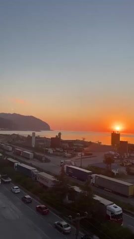 The video shows a sunrise over a coastal landscape. In the foreground, a road runs parallel to a body of water, with several cars and trucks on it. The trucks appear to be parked or moving slowly. Behind the road, there are several industrial buildings and structures, including what looks like a construction site with silos and cranes. The sun is low on the horizon, casting a warm orange glow over the water and sky. Mountains are visible in the distance. The overall scene suggests a port or industrial area at dawn.