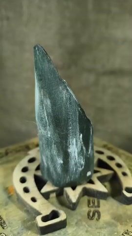 The video begins with a close-up shot of a dark, horn-shaped object standing upright on a wooden base. The object is rough and textured, with some white residue on its surface. The wooden base appears to be shaped like a horseshoe with a star inside. As the video progresses, the camera slowly pans around the horn-shaped object, revealing more of its surface. There are some lighter colored markings or engravings on the object, appearing gold or brass in color. The background is out of focus and appears to be a rough, textured surface, possibly a wall or fabric. The lighting is soft and diffused, casting subtle shadows. The video is short, lasting only a few seconds, and shows no people or discernible actions beyond the slow rotation of the camera.