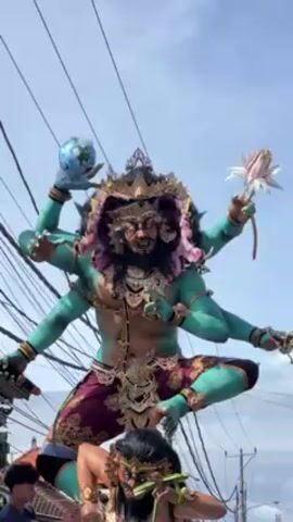 A large, elaborate effigy is being carried down a street by a group of people. The effigy is two-tiered. The top tier features a teal-skinned figure with six arms, wearing a crown and ornate jewelry, and holding a globe in one hand and a flower in another. The bottom tier shows a darker-skinned figure with a similar crown, holding green leaves in its mouth. People are walking alongside the effigy, guiding it through the street, which is lined with buildings and overhead power lines. The sky is partly cloudy. The scene appears to be a cultural event or procession.