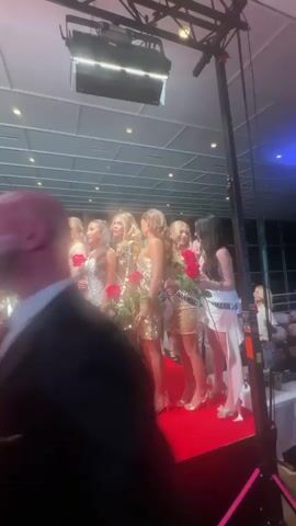 The video shows a group of women on a red carpet, likely at a beauty pageant event. The women are wearing similar dresses, some silver and some gold, and holding single red roses. They are standing in a row and posing for pictures. Many are smiling and looking at the camera. In the center is one woman wearing a red ballgown and a crown, holding a bouquet of roses. A photographer is taking pictures of them. The scene takes place indoors, likely in a conference center or event hall with a high ceiling.